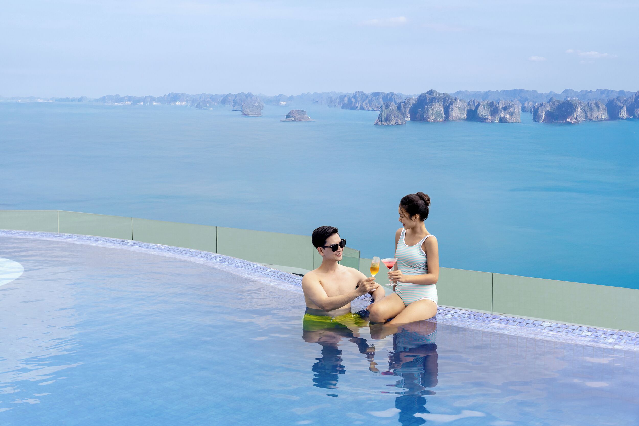 A couple sitting by an infinity pool with a view of the sea and rocky formations.