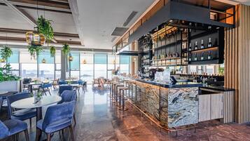 A modern bar area with a marble countertop, seating with bar stools, and a high ceiling with hanging plants.