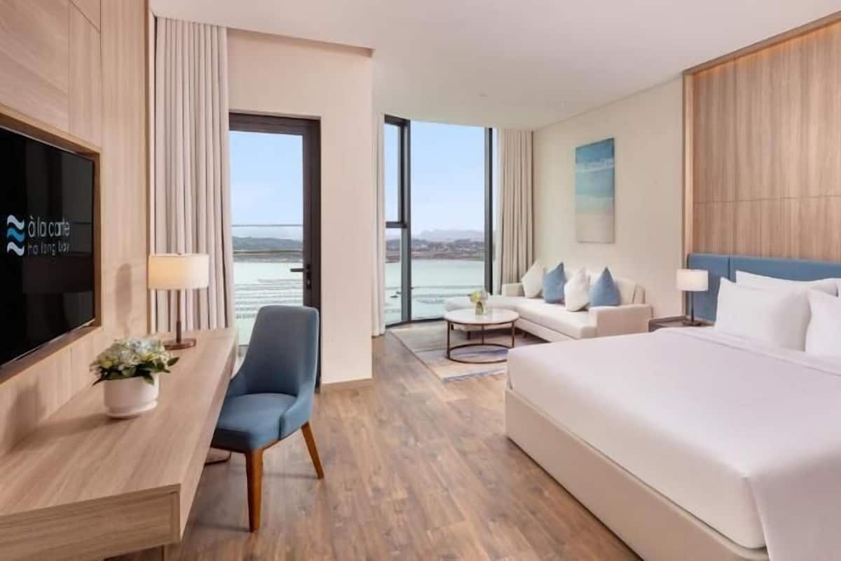 A hotel room with a bed, a desk, a chair, a sofa, a TV, and a view of the sea.