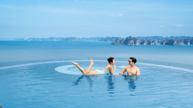 Una pareja relajándose en una piscina circular infinita con vistas a montañas distantes y al mar.