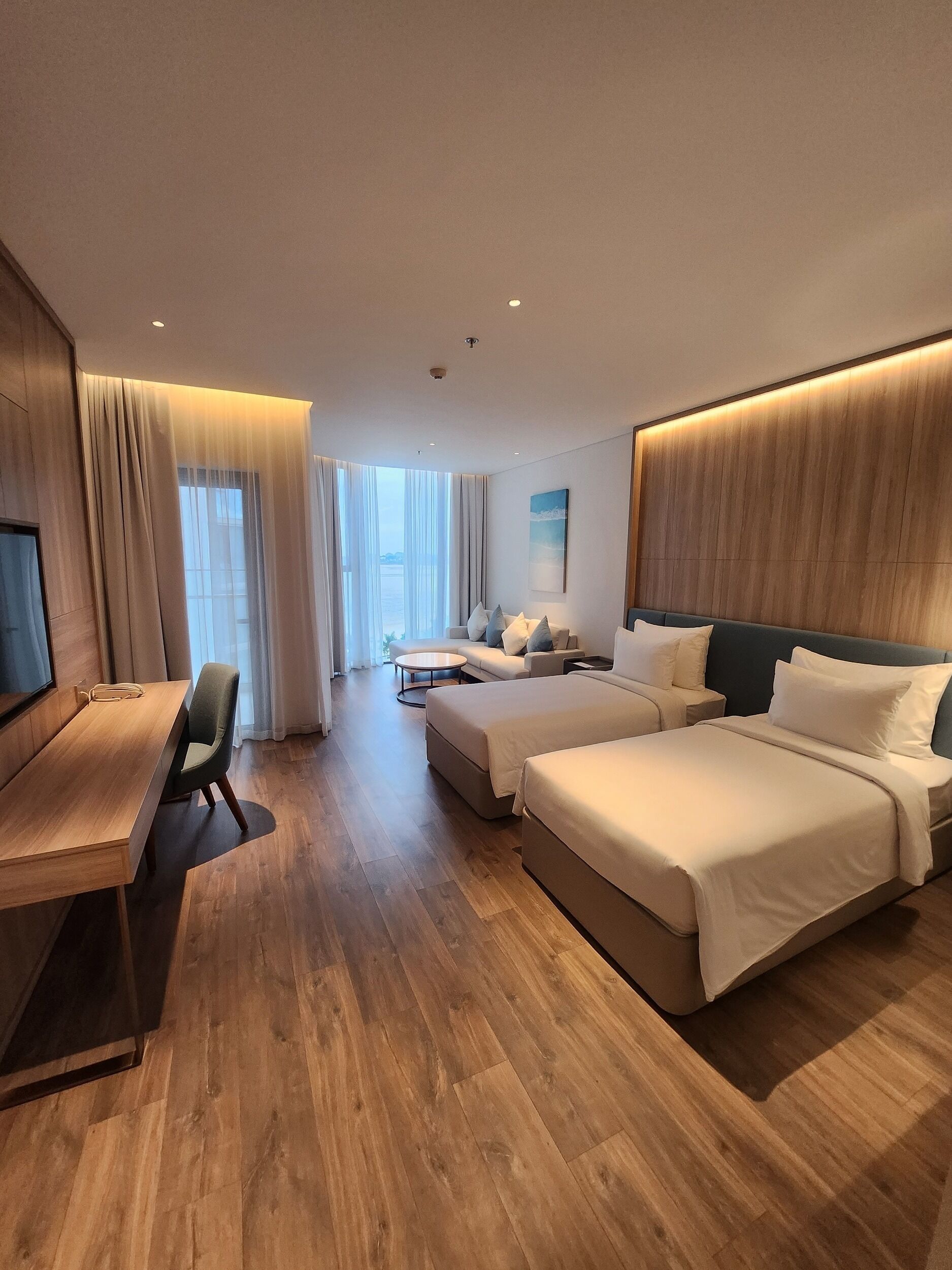 A hotel room with a wooden floor, a desk, two beds, a sofa, and a TV.
