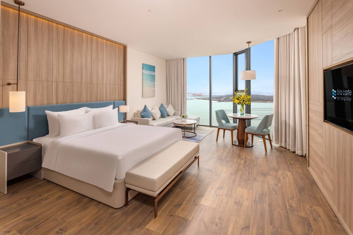 A modern hotel room with a large bed, a sofa, a dining table, and a TV.