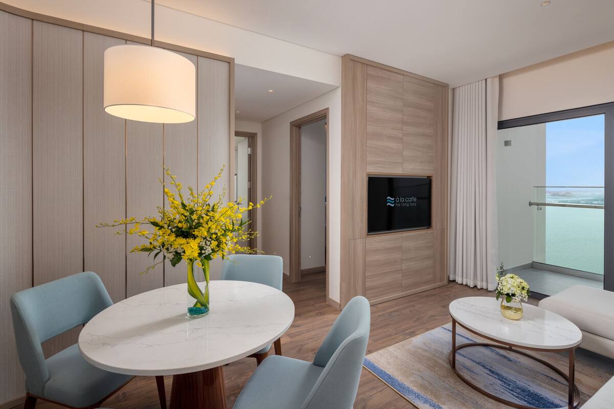 A modern hotel room with a round dining table, two chairs, a sofa, a TV mounted on the wall, and a large window offering a view.
