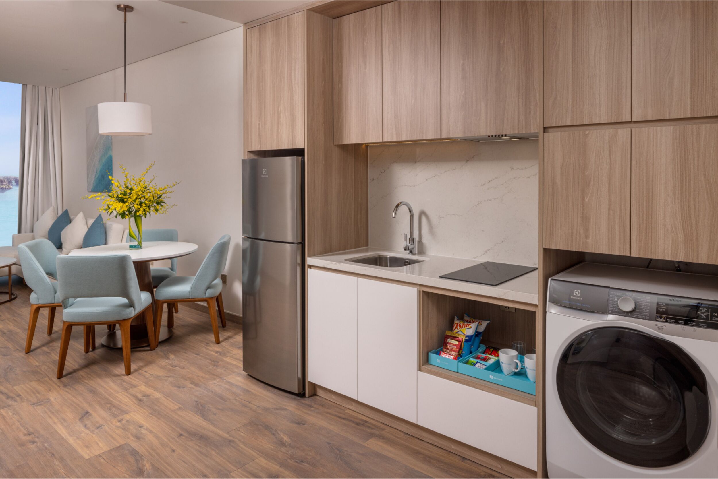 A modern kitchen with a built-in washing machine, a dining area with a round table and chairs, and a view of the sea through a large window.