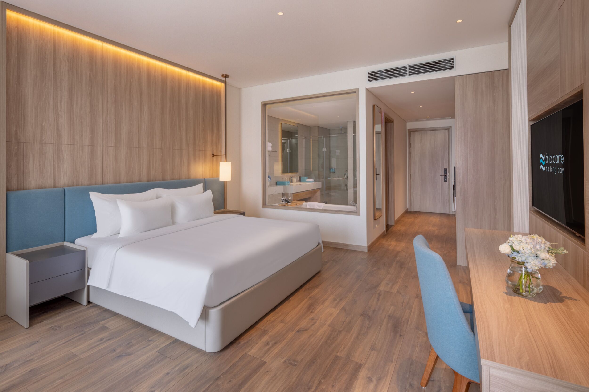 A modern hotel room with a large bed, a desk with a chair, a TV, and a bathroom visible through an open door.