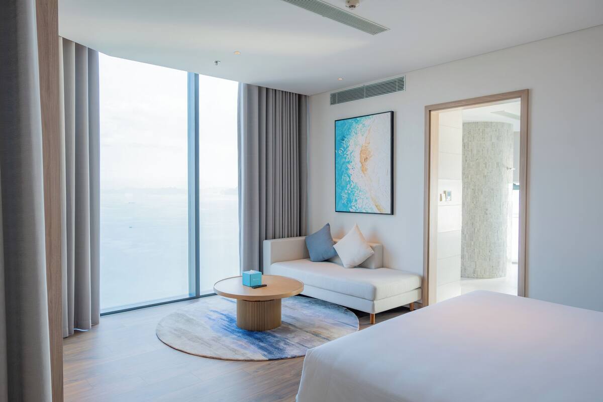 A modern hotel room with a large window, a round coffee table, a sofa, and a bed.