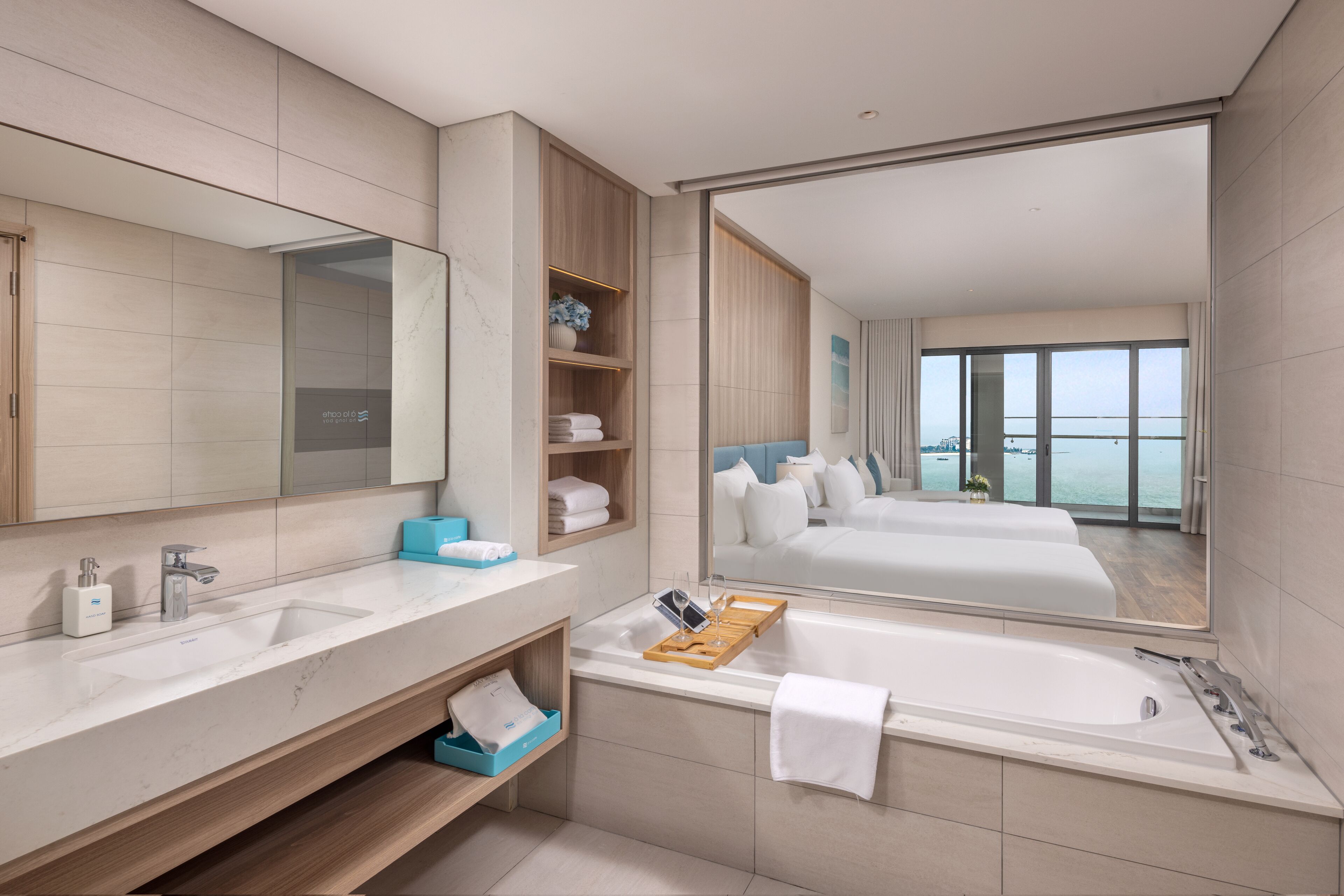 A modern bathroom with a large mirror, two sinks, a bathtub, and a view of a bedroom with a balcony.
