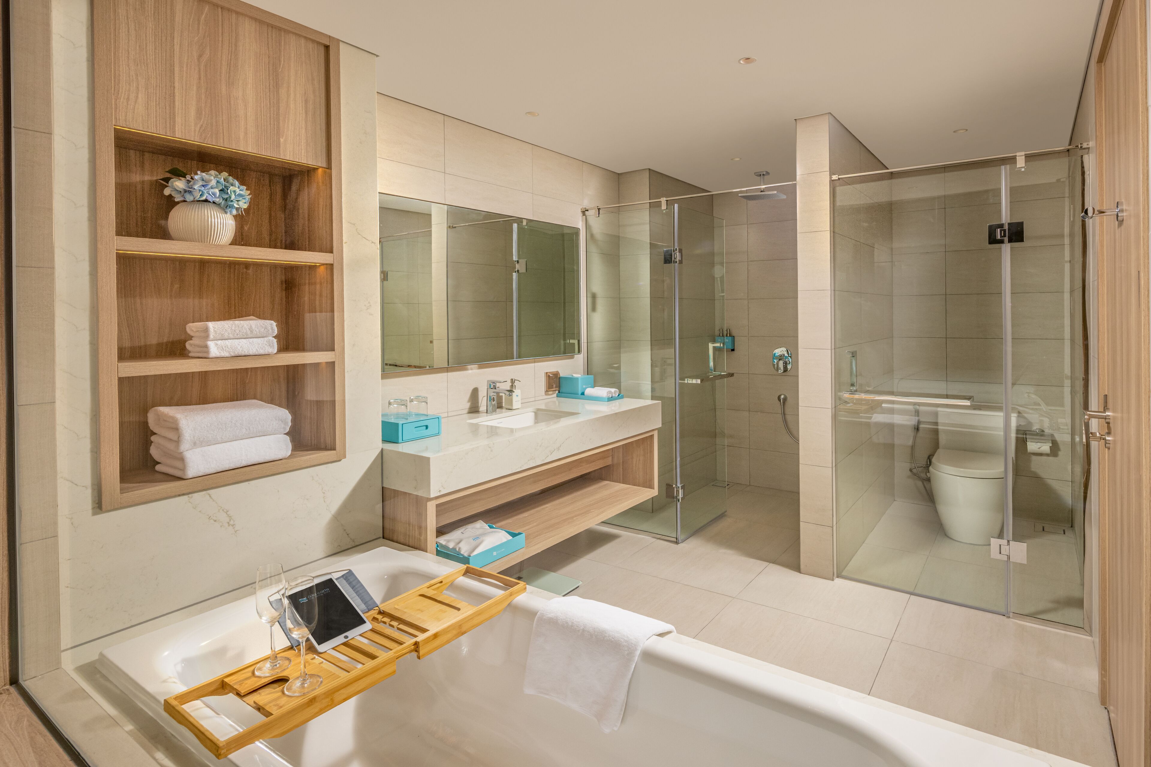 Un baño moderno con bañera, ducha con mampara de vidrio, tocador con espejo y repisas con toallas y artículos de aseo.
