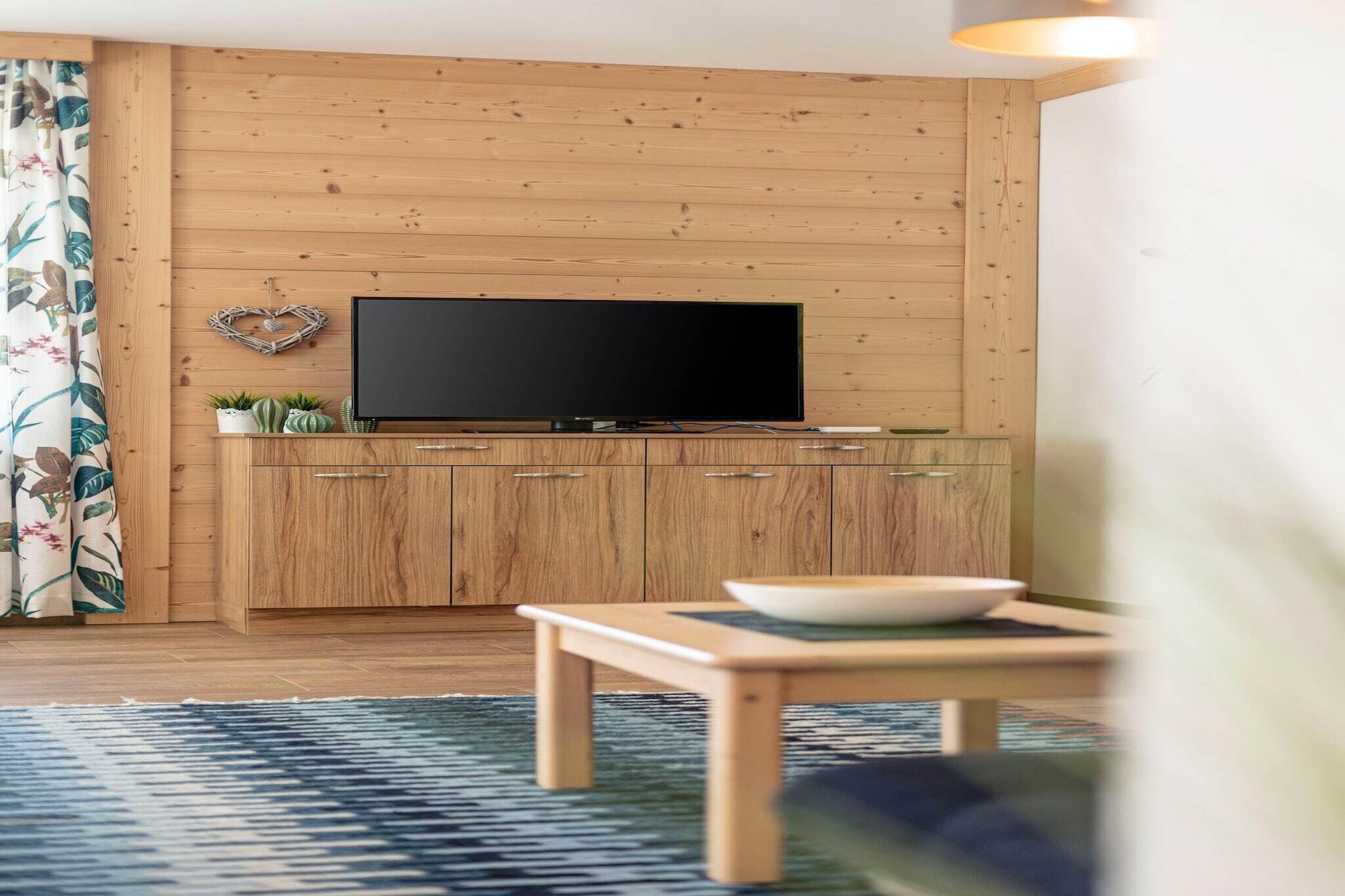 A living room with a wooden wall, a flat-screen TV mounted above a wooden cabinet, a small table with a bowl, and a patterned rug.