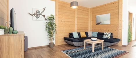 Ein modernes Wohnzimmer mit einer Holzverkleidung, einem schwarzen Sofa mit bunten Kissen, einem kleinen Holztisch und einer großen Topfpflanze.