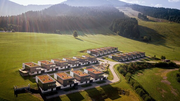 Eine Vogelperspektive auf einen modernen Wohnkomplex mit mehreren Gebäuden, grünen Rasenflächen und einer hügeligen Landschaft im Hintergrund.