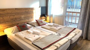 Habitación de hotel con cabecera de madera, dos camas con ropa de cama impecable, una lámpara de pared y vistas a un balcón a través de cortinas translúcidas.