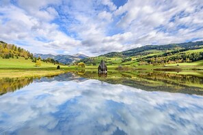 Un paisatge serè amb una casa al costat d'un estany tranquil, envoltat per turons suaus i un cel clar.