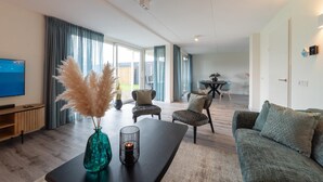 A modern living room with a flat-screen TV, a coffee table with decorative items, a sofa, and two armchairs. There is a dining area in the background with a table and chairs. - Holiday Home in Zeeland near the Sea (Colijnsplaat)