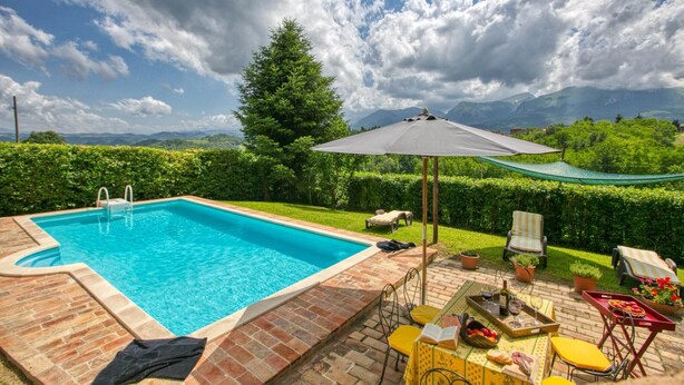 Piscina retangular com borda de tijolos, cercada por um espaço de área externa com cadeiras amarelas e vermelhas, uma mesa com bebidas e um guarda-sol verde.