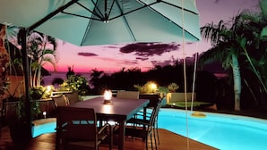 Ein Poolbereich mit Tisch und Stühlen, umgeben von Palmen und Blick auf einen Sonnenuntergang.