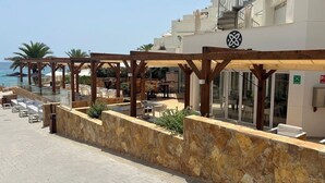 Un hotel frente a la playa con mesas al aire libre, paredes de piedra y vista despejada al mar.