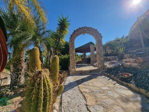 A stone pathway leading to a covered area with a stone archway, surrounded by cacti and palm trees. - Villa with private pool, large garden and incredible sea view! (El Campello)