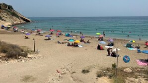 Una playa con gente tomando el sol y nadando, sombrillas de colores y un cielo despejado.