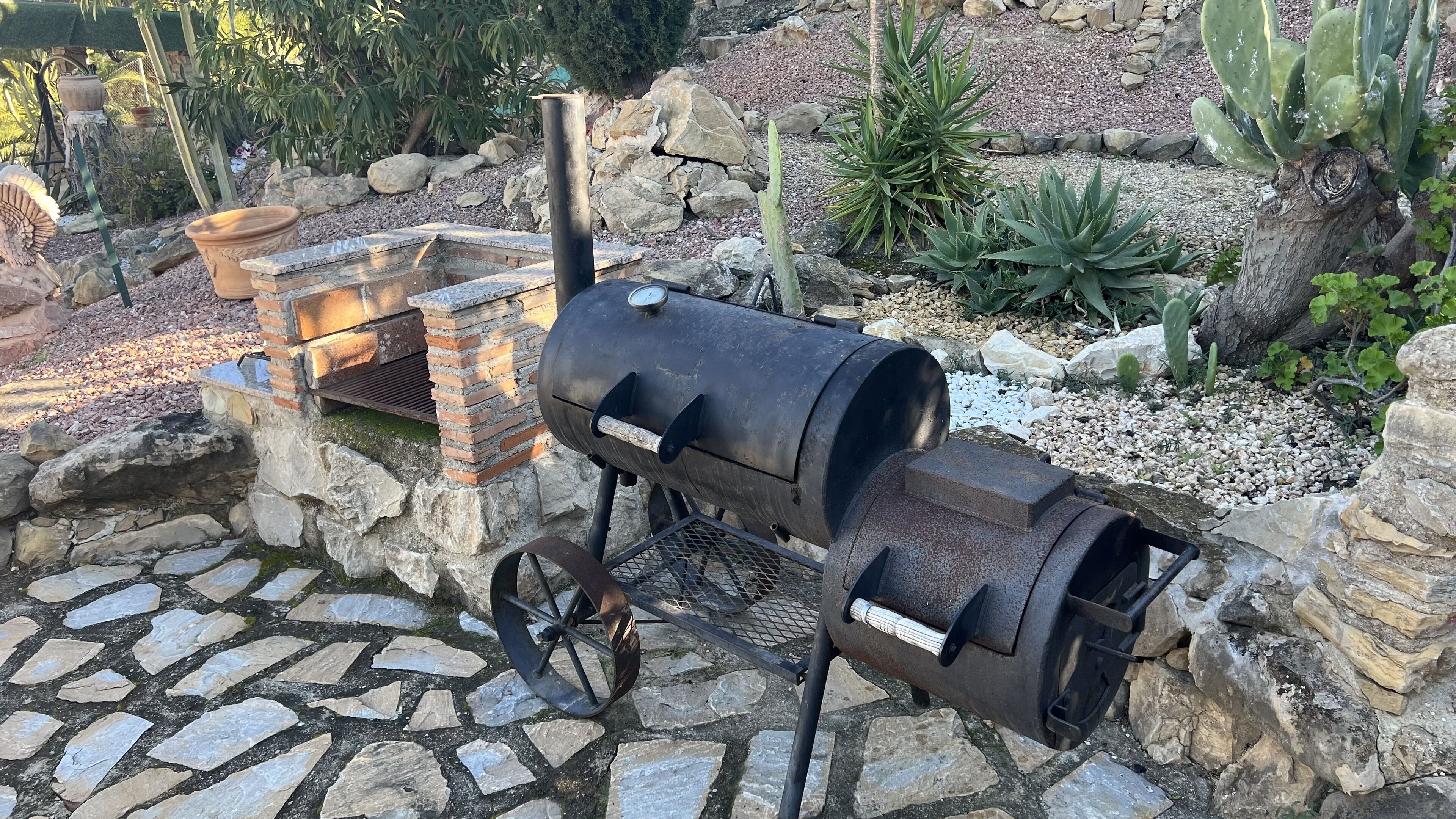 Un barbecue au charbon de bois doté d’une cheminée, installé sur une terrasse en pierre entourée de cactus et de rochers.
