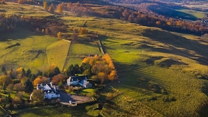 Luchtbeeld van een landelijk gebied met glooiende heuvels, een kleine groep huizen en een dicht bos op de achtergrond.