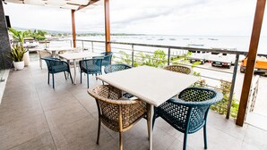 Uma área de estar coberta ao ar livre, com cadeiras de vime e uma mesa, com vista para um corpo d'água com barcos.