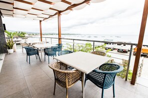 A covered outdoor seating area with wicker chairs and a table, overlooking a body of water with boats. - Hotel Dulcee (Naga City)