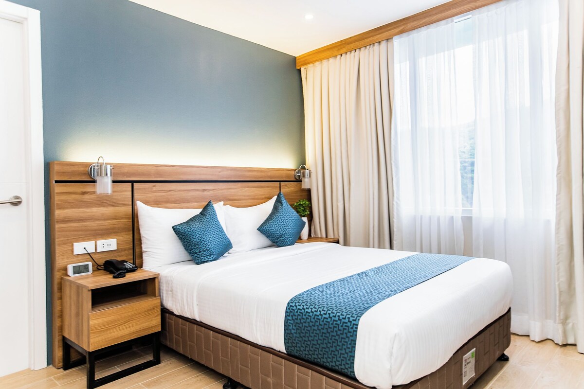 A hotel room with a bed, bedside table, lamp, and curtains.