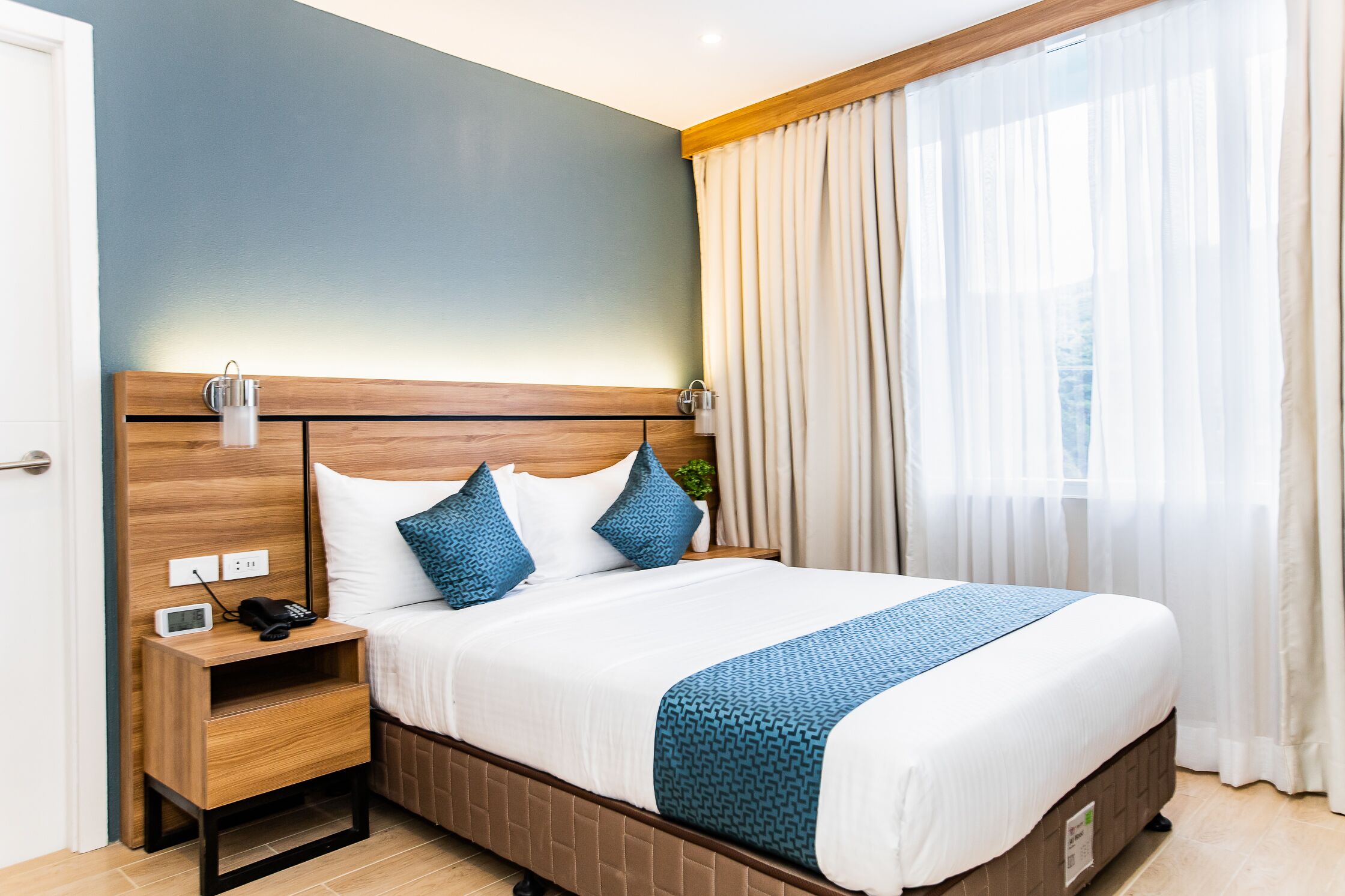 A neatly made bed with a blue blanket, a bedside table with a phone, and a nightstand with a lamp.