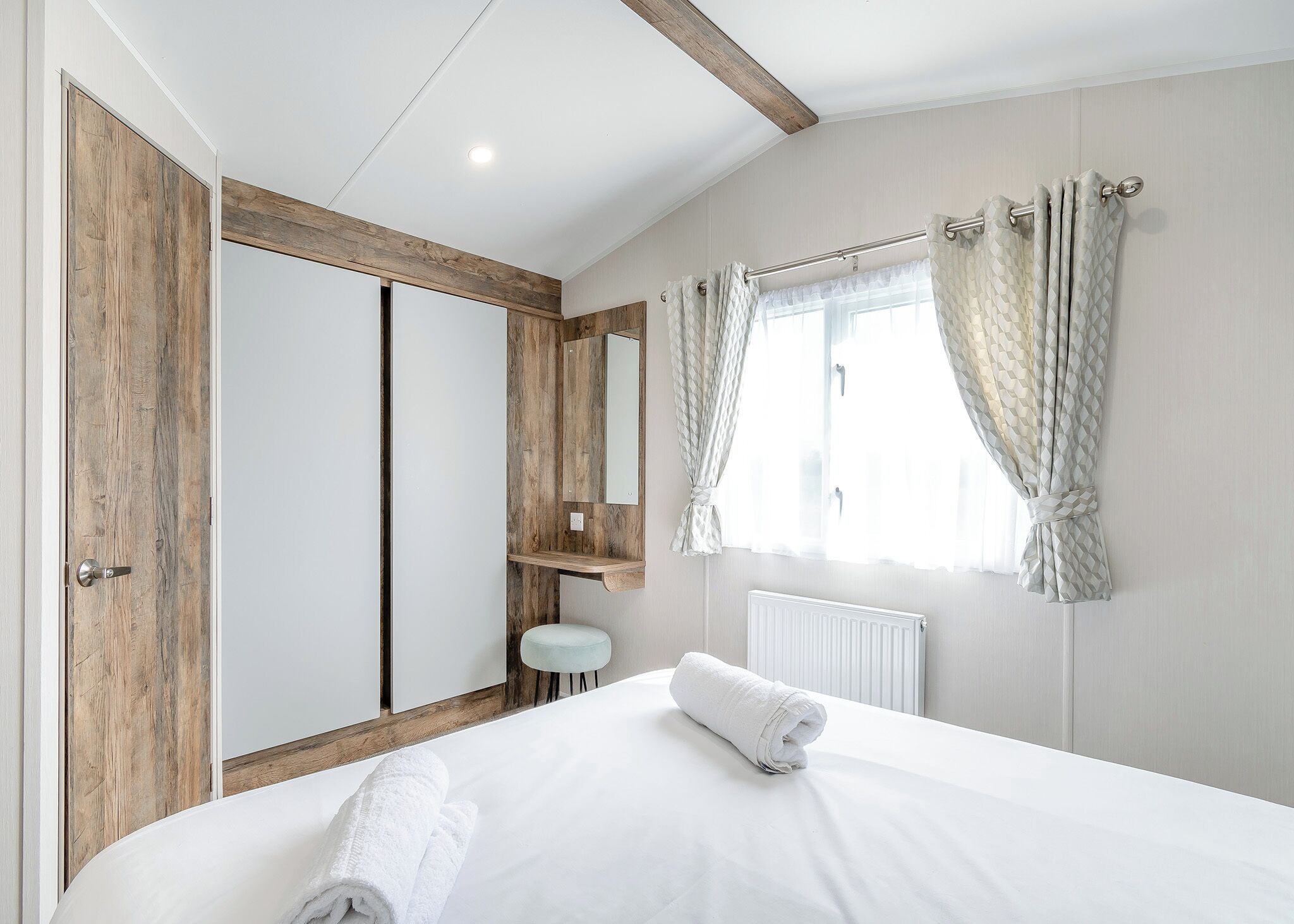 Un dormitorio con cama, armario de madera, espejo, radiador y ventana con cortinas.