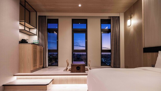 A modern hotel room with a large bed, a desk with a computer, and a view of the city at night through three large windows.