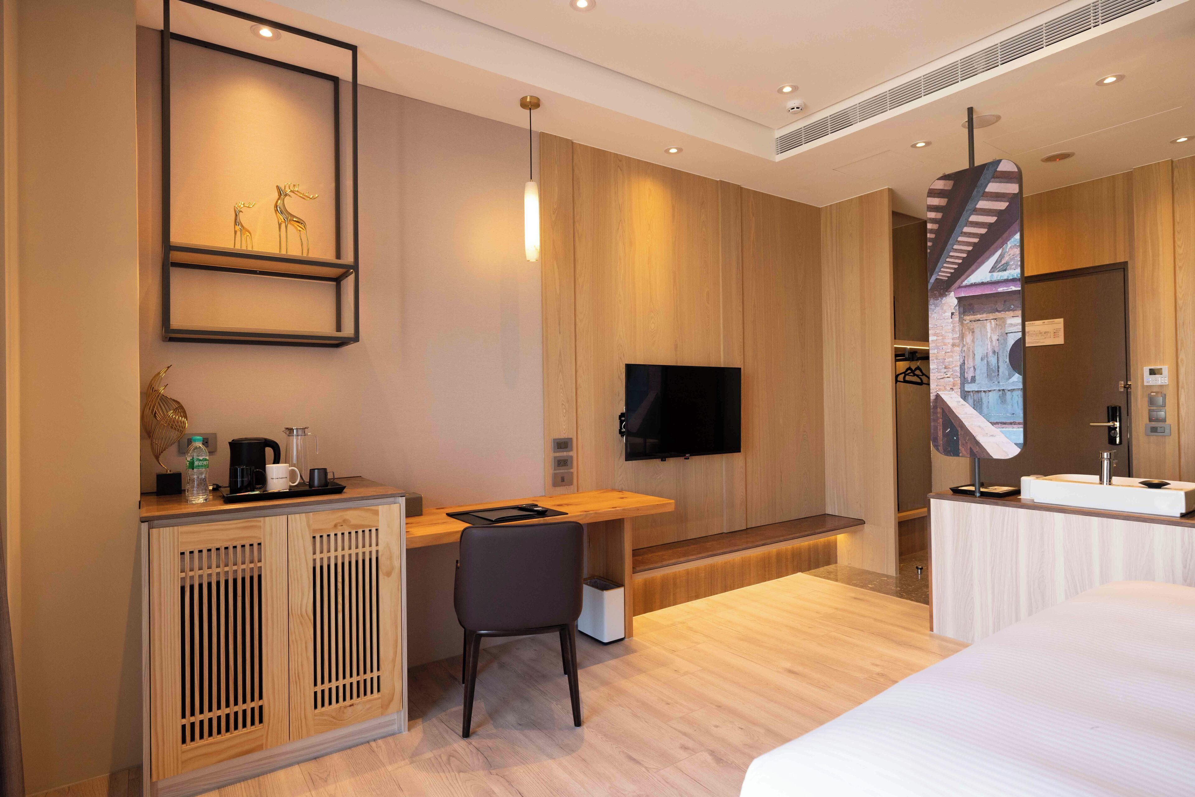 A modern hotel room with a wooden desk, a flat-screen TV, a bed, and a bathroom with a mirror and sink.