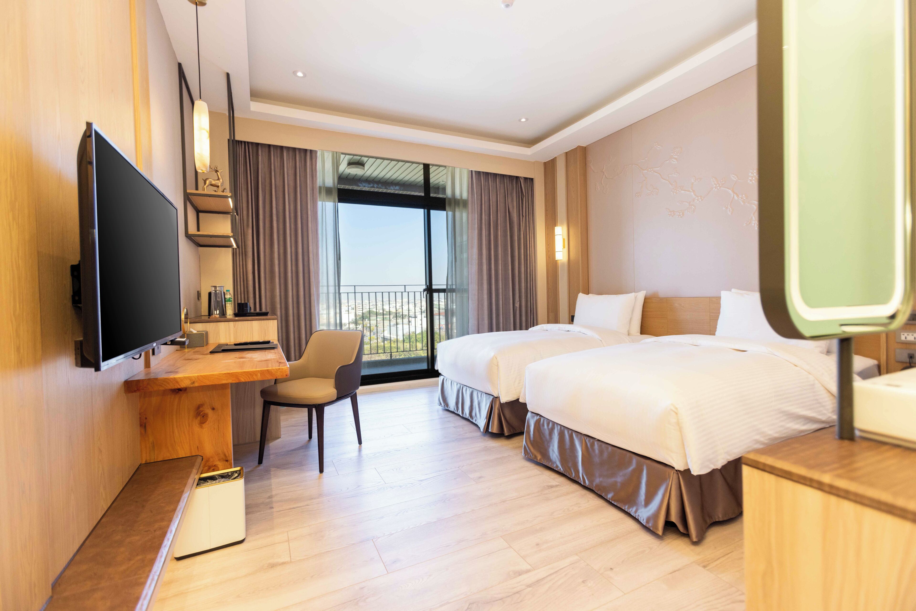 A hotel room with a large flat-screen TV, a desk with a chair, two beds, and a balcony with curtains.