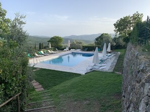 A rectangular swimming pool with lounge chairs and umbrellas, surrounded by a stone wall and a grassy area with a view of distant hills.