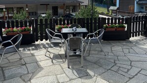 Um pátio externo com piso de pedra, cadeiras de metal e uma mesa com guarda-sol verde.