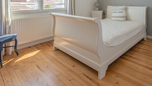 Een slaapkamer met een wit bed, een nachtkastje, een lamp en uitzicht op buiten door een raam.