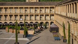 Un cortile con un'alta torre campanaria, circondato da un edificio con più archi e un porticato.
