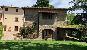 A stone building with a tiled roof, a small arched entrance, and a flower bed with red flowers.