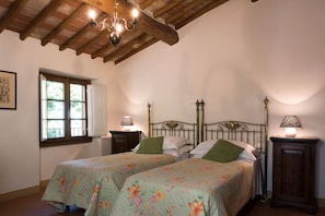 A bedroom with two beds, a wooden ceiling, a window with shutters, and bedside lamps.