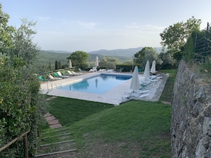 A rectangular swimming pool with lounge chairs and umbrellas, surrounded by a stone wall and a grassy area with a view of distant hills.