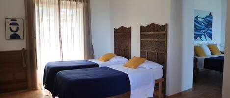 A hotel room with two beds, a wooden headboard, a large window with curtains, and a painting on the wall.