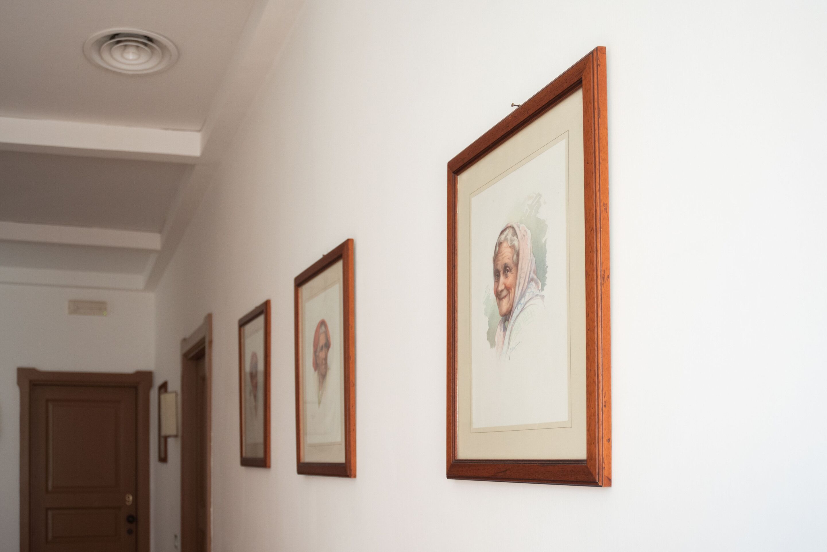 A hallway with framed portraits on the wall, a wooden door, and a ceiling light.