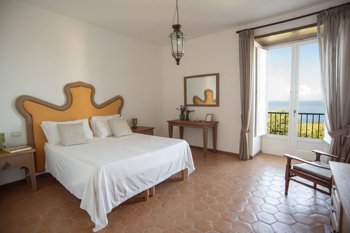 Une chambre à coucher avec un grand lit, une table de chevet, une commode, une chaise, une petite table avec un vase, un miroir et une vue sur la mer par une fenêtre avec des rideaux.