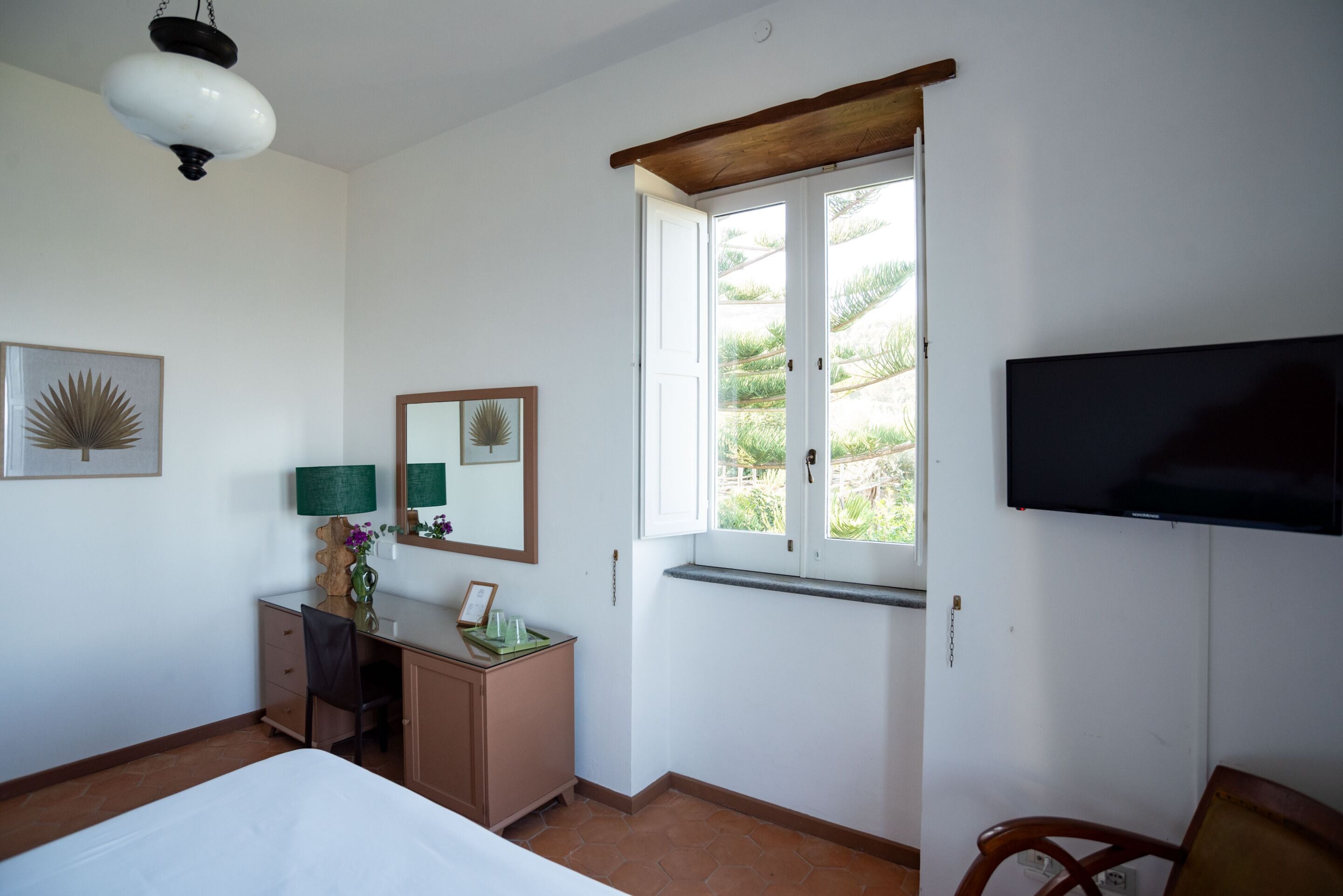 Habitación de hotel con cama, escritorio con silla, televisor, ventana con vistas a zonas verdes y una lámpara en el techo.