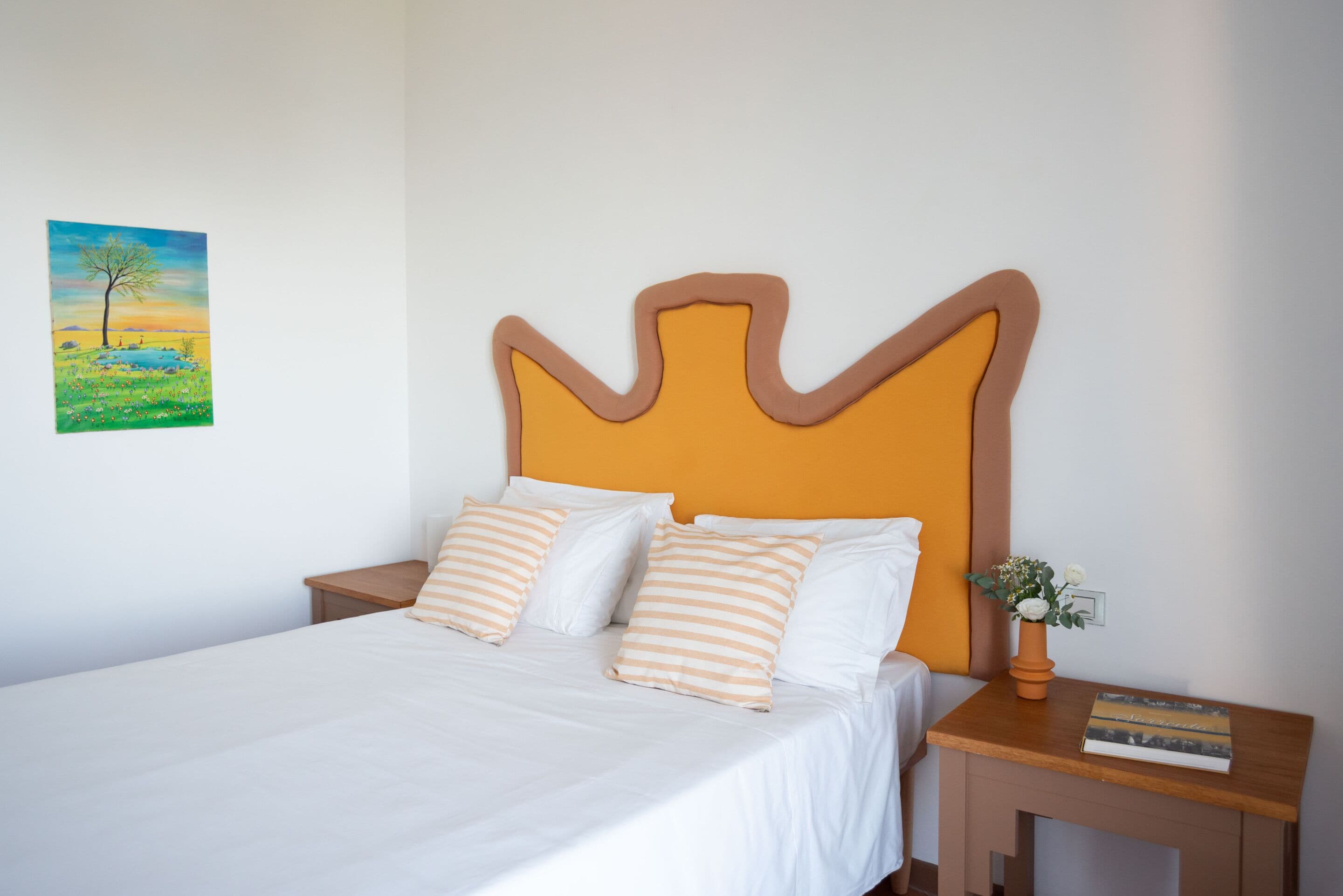 A bed with white bedding and striped pillows, a wooden nightstand with a book and a small plant, and a painting of a landscape on the wall.
