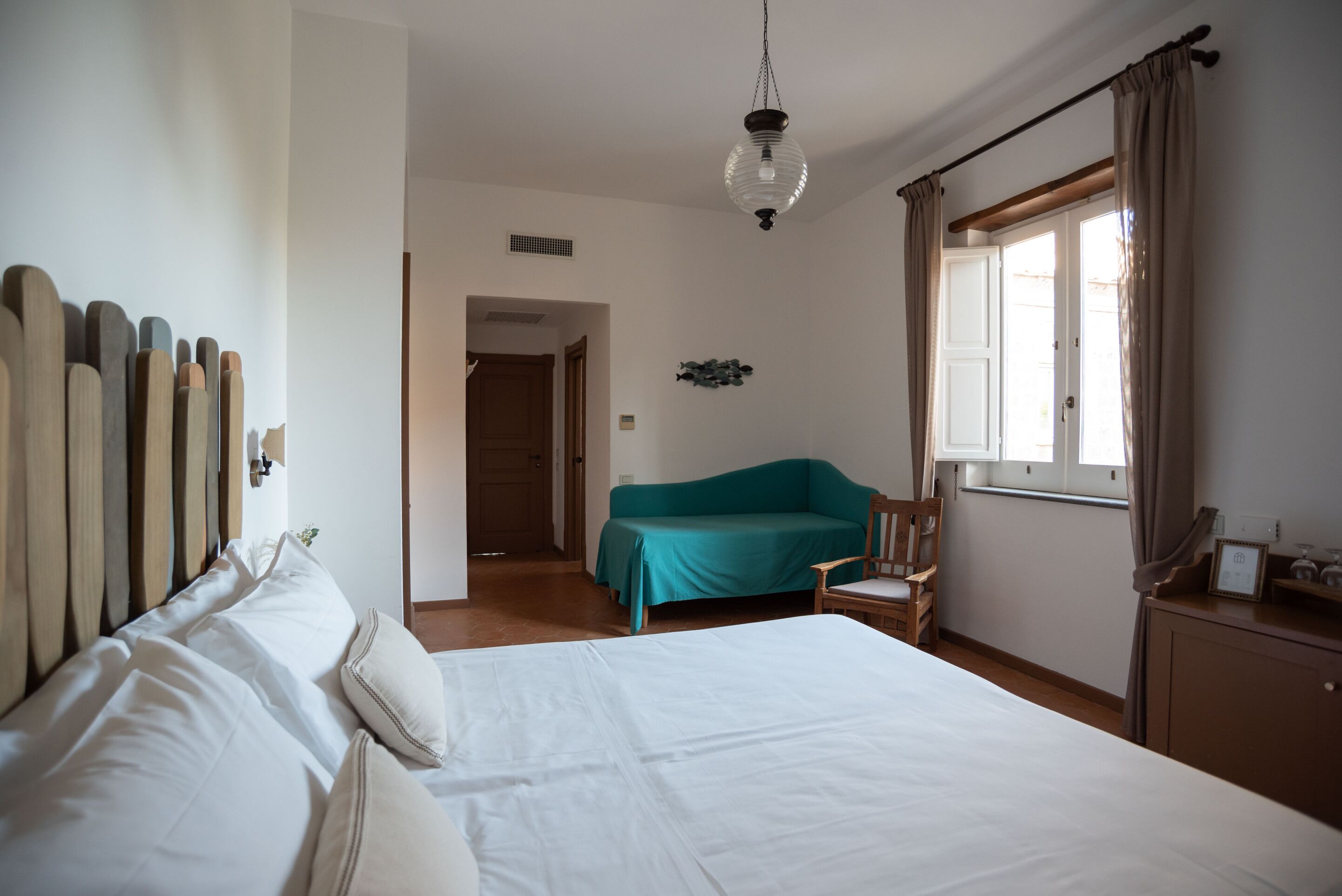 A hotel room with a large bed, a teal sofa, a wooden chair, and a window with curtains.