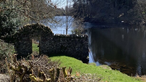 Uma ponte de pedra sobre um rio calmo, cercada por árvores e vegetação.