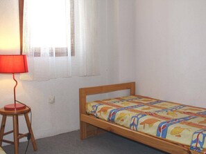 A small, single-bed room with a wooden frame and a colorful bedspread. There is a red lamp on a wooden stool and a white lace curtain on the window. - Charming 2BR Pavilion with Pool Access, Terrace, and Parking, 400m from Sea (Gruissan)