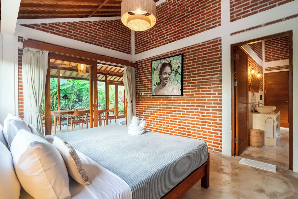 A bedroom with a brick wall, a bed with white bedding, a wooden ceiling, and a large window with curtains.