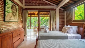 A room with a brick wall, a bed with white bedding, a wooden dresser, and a large window with curtains. - Kampung Uma Dawa Villas and Spa (Ubud)